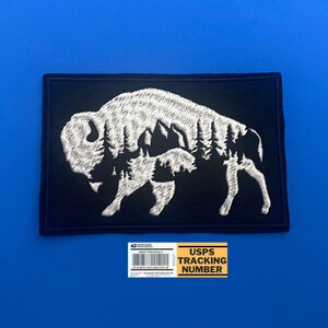 May include: Black embroidered patch with a white silhouette of a bison with trees inside its outline. The patch is square shaped and has a white border.