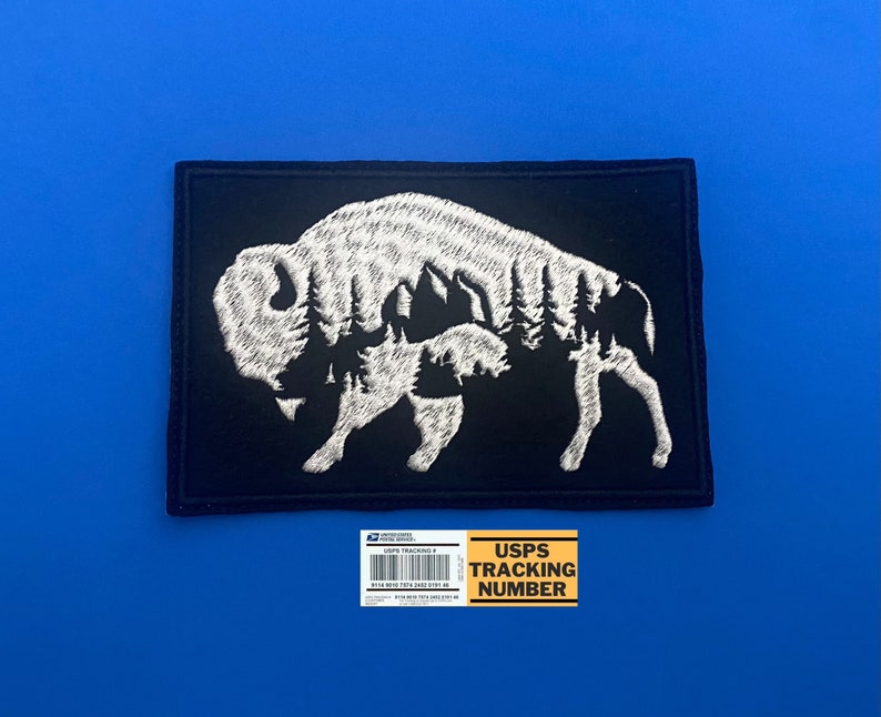 May include: Black embroidered patch with a white silhouette of a bison with trees inside its outline. The patch is square shaped and has a white border.