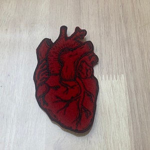May include: A red felt anatomical heart patch with black embroidery. The heart is detailed with veins and chambers.