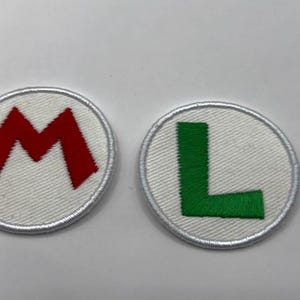 May include: Two circular embroidered patches with white backgrounds and white stitched borders. One patch features a bold red letter M, and the other patch features a bold green letter L, both rendered in a block font style.