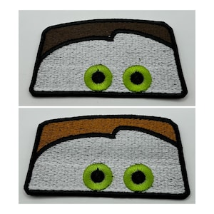 May include: Two rectangular embroidered patches featuring a stylized character design. Each patch displays a white face area with two bright green eyes with black pupils, topped by a brown hair section, all outlined in a thick black border.