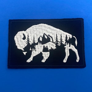 May include: White embroidered patch of a bison silhouette with a forest scene inside on a black background.