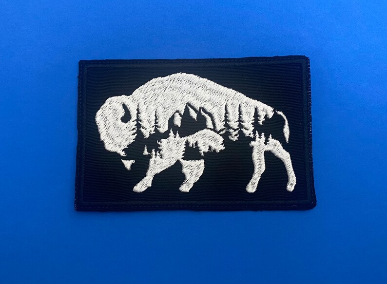 May include: White embroidered patch of a bison silhouette with a forest scene inside on a black background.