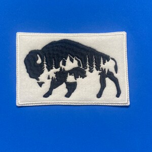 May include: White embroidered patch with a black silhouette of a buffalo with a forest inside its shape.
