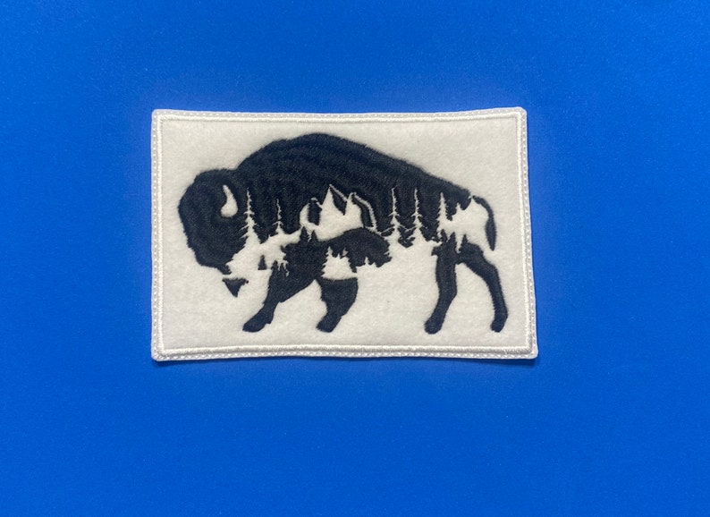 May include: White embroidered patch with a black silhouette of a buffalo with a forest inside its shape.