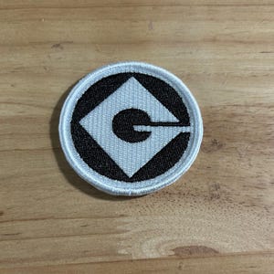 May include: A white and black embroidered patch with the Minions logo. The logo is a white diamond shape with a black 'G' inside.