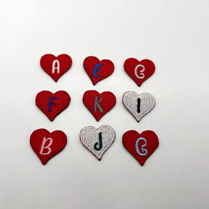 May include: Nine heart-shaped embroidered patches. Each patch is red, with a white or blue embroidered letter. The letters are A, E, G, F, K, I, B, J, and G. The patches are arranged in a three-by-three grid.