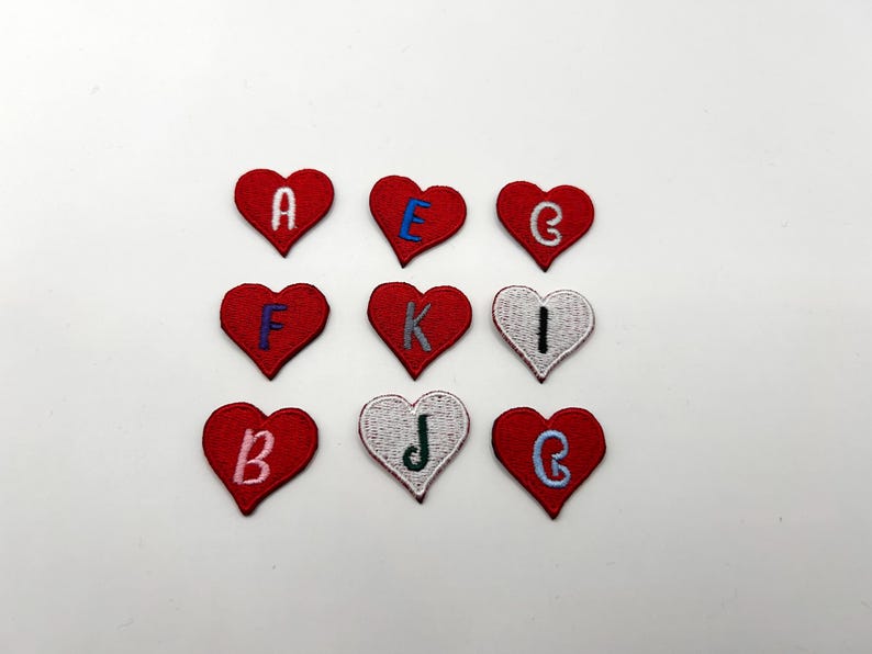 May include: Nine heart-shaped embroidered patches. Each patch is red, with a white or blue embroidered letter. The letters are A, E, G, F, K, I, B, J, and G. The patches are arranged in a three-by-three grid.