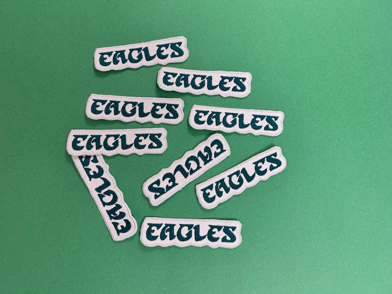 May include: A collection of nine white fabric patches with the word "Eagles" written in a green cursive font. The patches are arranged in a random pattern on a green background.