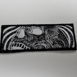 May include: Black and white embroidered patch with a skull design. The skull is surrounded by a wavy pattern.