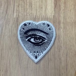 May include: Heart-shaped embroidered patch featuring a detailed eye design in black and white. The eye is surrounded by radiating lines and small stars, with teardrop-like shapes at the bottom. The patch is on a wooden surface.