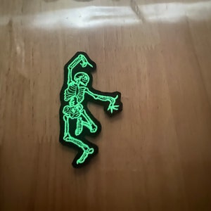 Puede incluir: Un parche negro con un esqueleto verde brillante en una pose de baile. Los huesos del esqueleto est&aacute;n detallados y el parche tiene un borde negro. Adecuado para ropa o accesorios.