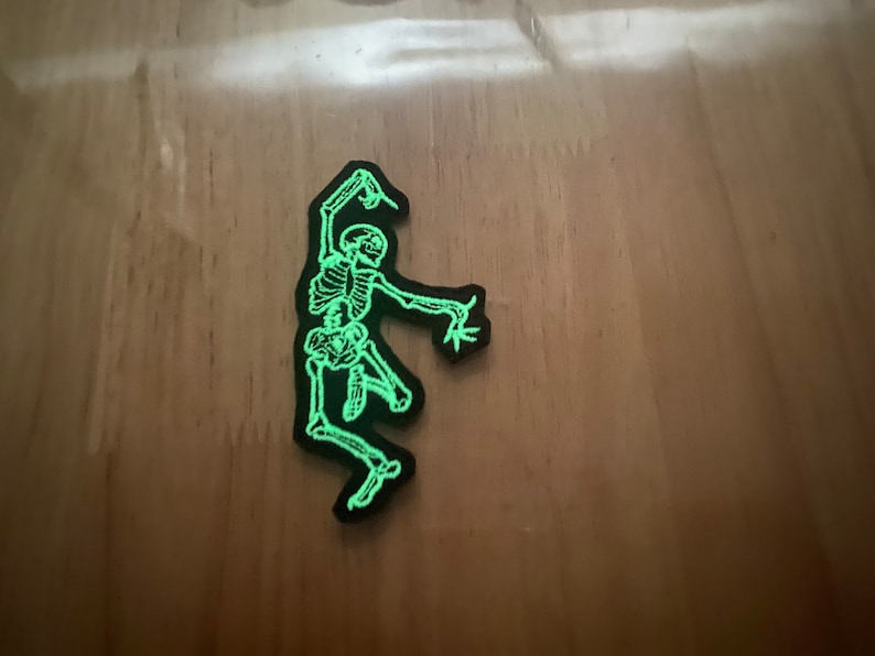 Puede incluir: Un parche negro con un esqueleto verde brillante en una pose de baile. Los huesos del esqueleto est&aacute;n detallados y el parche tiene un borde negro. Adecuado para ropa o accesorios.