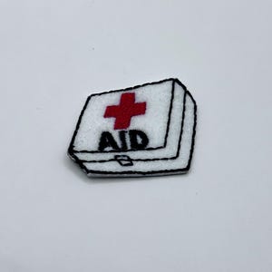 May include: A white felt patch in the shape of a first aid kit. The patch features a red cross and the word "AID" in black letters. The edges are outlined in black.