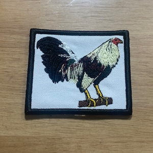 May include: Embroidered patch featuring a white and black rooster with a red comb, standing on a brown branch. The patch has a black border and is on a white background.