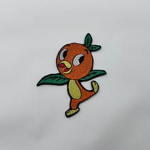 May include: Embroidered patch featuring an orange bird character with green leaf accents. The bird has a yellow belly, black eyes, and is posed with its arms and one leg extended. The patch is designed for application on clothing or accessories.