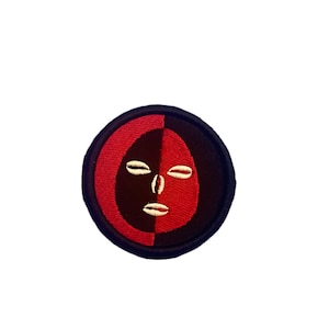 May include: A circular embroidered patch with a black and red background. The patch features a stylized face with white eyes and a mouth.