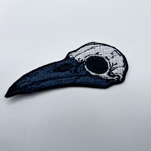 May include: Embroidered patch featuring a stylized plague doctor skull. The patch has a white skull and a dark blue beak. The eye socket is a large, dark circle. The patch is on a white background.