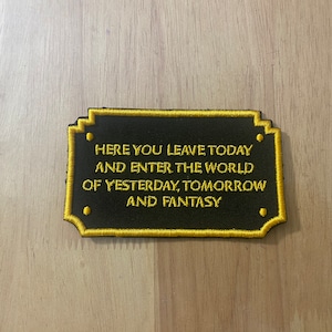 May include: A black embroidered patch with gold stitching. The patch features the text "Here you leave today and enter the world of yesterday, tomorrow and fantasy."