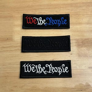 May include: Three black fabric patches with white embroidery that reads "We the People". The top patch has red and blue embroidery. The patches are on a wood surface.