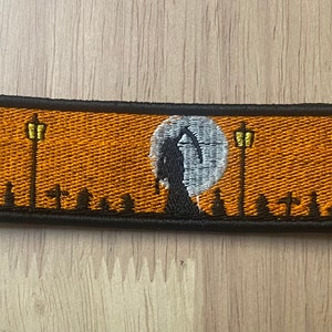 May include: An embroidered patch with a black silhouette of the Grim Reaper standing in front of a white moon. The background is orange with two yellow streetlights and black silhouettes of tombstones.