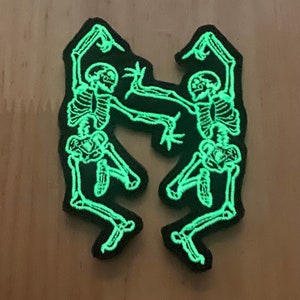 Puede incluir: Dos parches negros con un dise&ntilde;o de esqueleto verde fosforescente. Los esqueletos est&aacute;n bailando, con los brazos y las piernas extendidos. Los parches est&aacute;n sobre una superficie marr&oacute;n claro.