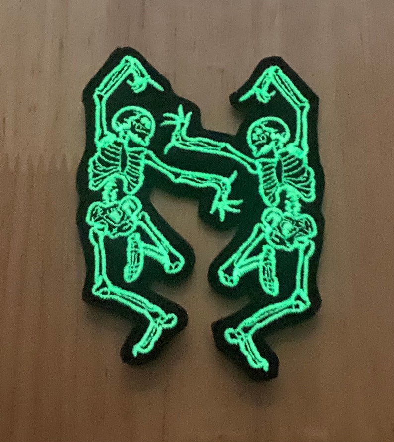 Puede incluir: Dos parches negros con un dise&ntilde;o de esqueleto verde fosforescente. Los esqueletos est&aacute;n bailando, con los brazos y las piernas extendidos. Los parches est&aacute;n sobre una superficie marr&oacute;n claro.