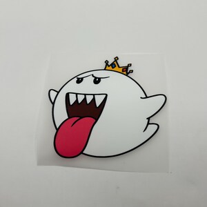 May include: A white ghost sticker with a round body, small arms, and a wide open mouth showing sharp teeth and a long pink tongue. The ghost has a yellow crown with blue and red accents on its head and is outlined in thick black lines.