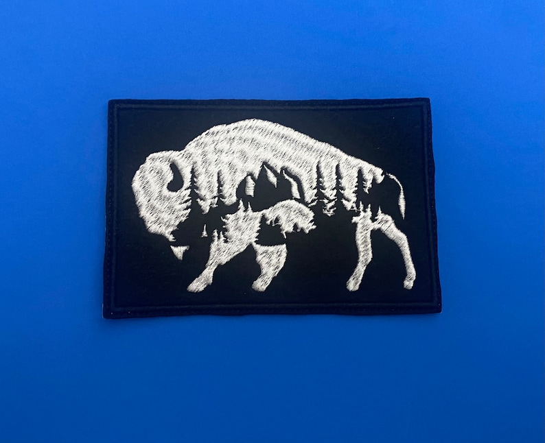 May include: White embroidered patch of a bison with a forest inside its silhouette on a black background.