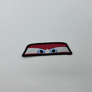 May include: Embroidered patch with a design of blue eyes peeking out from behind a red and white striped shape, outlined in black. A playful, whimsical accessory for clothing or bags.