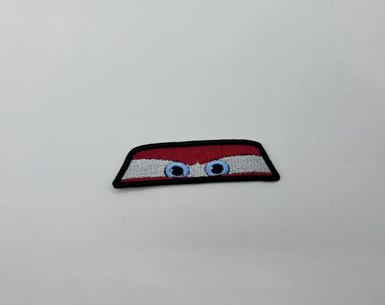 May include: Embroidered patch with a design of blue eyes peeking out from behind a red and white striped shape, outlined in black. A playful, whimsical accessory for clothing or bags.