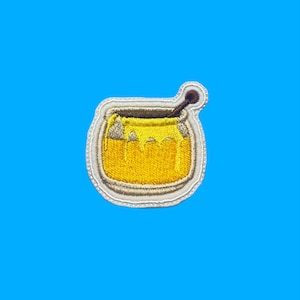 May include: Embroidered patch featuring a yellow pot of honey with a wooden honey dipper. The patch has a white border and is set against a bright blue background. The honey appears to be dripping from the pot.