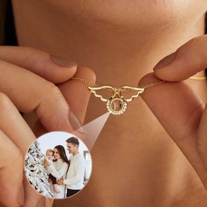 Può includere: Una collana color oro con un ciondolo raffigurante una foto di famiglia. Il ciondolo è circondato da piccole pietre trasparenti e ha un design ad ali. La collana è tenuta da una persona.