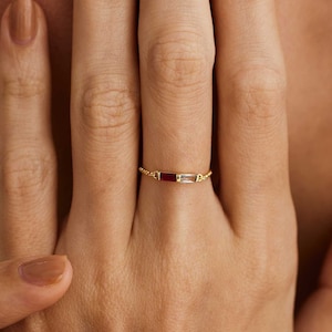 May include: A delicate gold chain ring with a red and clear baguette-shaped gemstone setting.