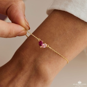 May include: A gold chain bracelet with a pink and red heart charm. The charm is made of two heart-shaped stones, one pink and one red.