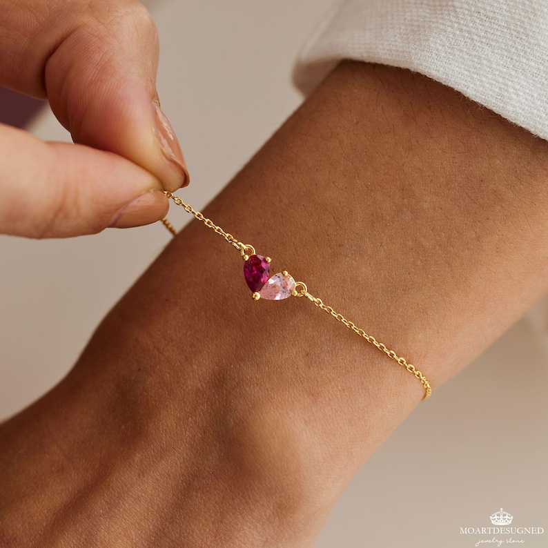 May include: A gold chain bracelet with a pink and red heart charm. The charm is made of two heart-shaped stones, one pink and one red.