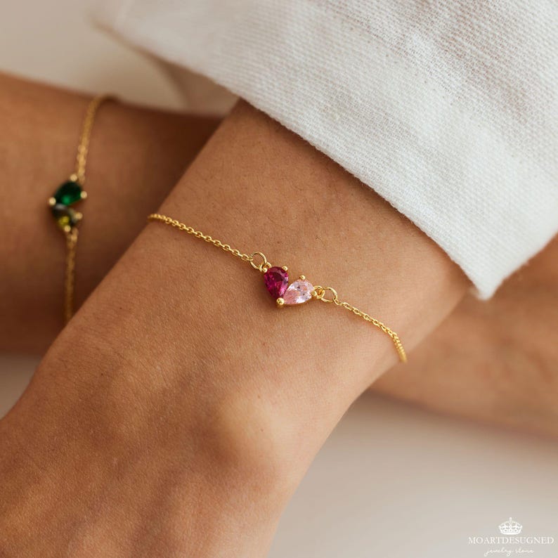 May include: A gold chain bracelet with a pink and red heart-shaped charm. The charm is made of two teardrop-shaped stones.