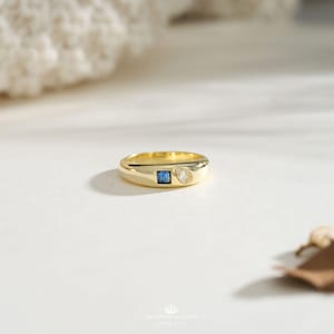 May include: A gold band ring featuring a small square-cut blue gemstone and a teardrop-shaped clear gemstone set side-by-side on the top surface. The ring has a polished, smooth finish and a rounded profile, showcasing a clean and simple design.