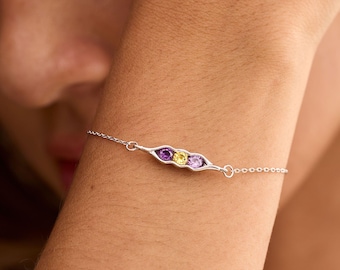 May include: A silver chain bracelet featuring a pea pod-shaped charm containing three round faceted gemstones: a purple amethyst, a yellow citrine, and a purple amethyst. The metal charm is polished silver with a delicate link chain.