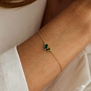 May include: A gold chain bracelet with two green gemstone charms. The charms are in the shape of teardrops and are set in gold.