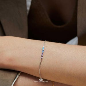 May include: A delicate silver bracelet featuring a row of rectangular, multi-colored gemstones in shades of blue and purple. The bracelet is adorned with a small crown charm. The text "MOARTDESIGNED" is visible.
