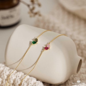 May include: Two delicate gold chain bracelets with heart-shaped charms. One charm features green and brown gemstones, while the other has pink and red gemstones.
