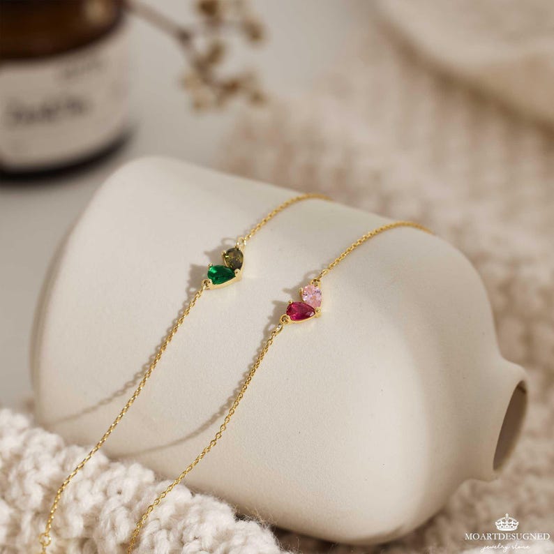 May include: Two delicate gold chain bracelets with heart-shaped charms. One charm features green and brown gemstones, while the other has pink and red gemstones.