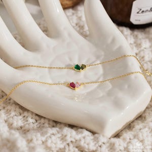 May include: Two delicate gold chains with heart-shaped pendants. The top chain features a green and brown heart, while the bottom chain has a pink and red heart.