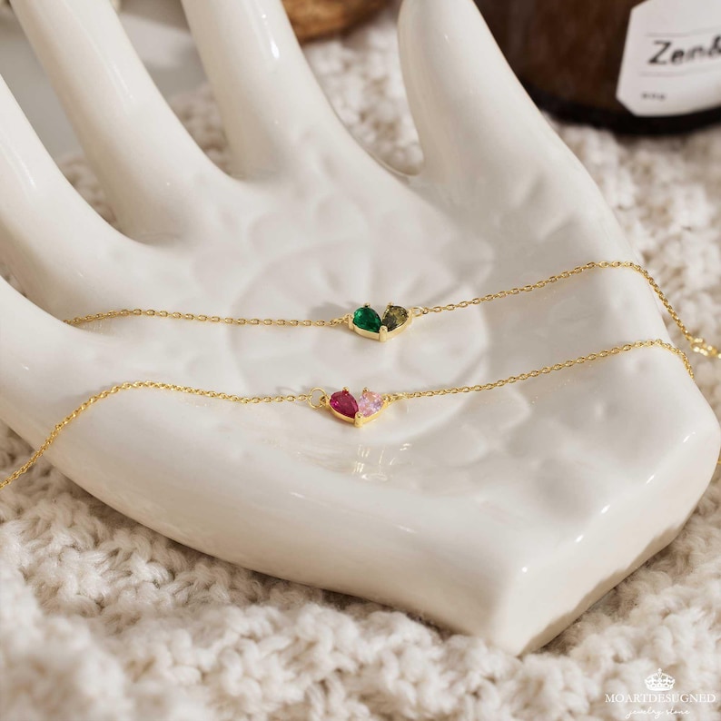 May include: Two delicate gold chains with heart-shaped pendants. The top chain features a green and brown heart, while the bottom chain has a pink and red heart.