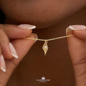 May include: A gold necklace featuring a delicate chain and a pendant shaped like a stylized pod or eye, containing a single small white pearl at its center. The pendant has a smooth, polished finish and a pointed bottom, with the text MOARTDESUGNED.