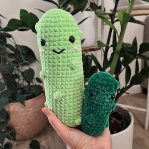 May include: Two crocheted pickle-shaped plush toys. The larger one is light green with black eyes and a smiling face with pink cheeks. The smaller one is dark green. Both are held in a hand.