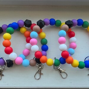 May include: Three colourful beaded keyrings with black accents and silver key rings. Each keyring is made of round silicone beads in a rainbow pattern.