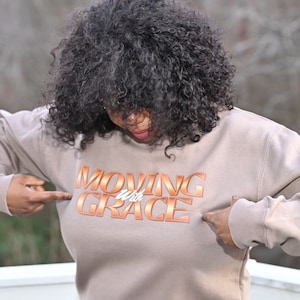 May include: A light brown sweatshirt with the text "MOVING with GRACE" in a copper-colored font.