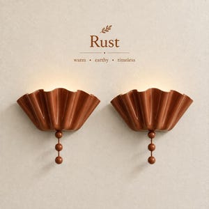 May include: Two rust-colored wall sconces with a pleated, fan-like shade and a vertical stem featuring three spherical beads. The text above reads Rust, warm, earthy, timeless. The fixtures emit a soft, warm glow from the top of the pleated shades.